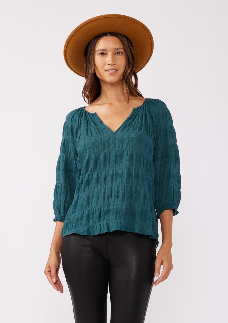 [Color: Deep Teal] A front facing image of a brunette model wearing a textured jacquard stripe blouse in teal. A relaxed fit blouse with a split v neckline and long sleeves. Styled with dark denim for the fall season. 