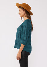 [Color: Deep Teal] A side facing image of a brunette model wearing a textured jacquard stripe blouse in teal. A relaxed fit blouse with a split v neckline and long sleeves. Styled with dark denim for the fall season. 