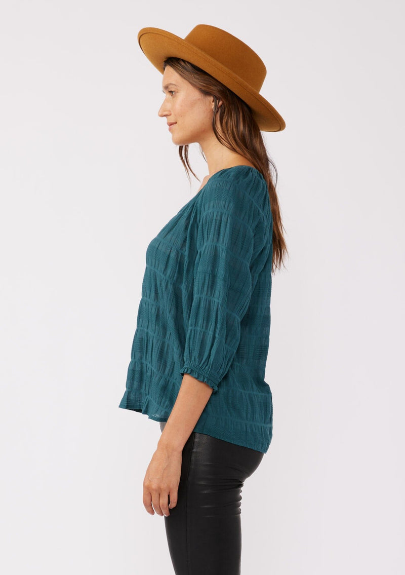 [Color: Deep Teal] A side facing image of a brunette model wearing a textured jacquard stripe blouse in teal. A relaxed fit blouse with a split v neckline and long sleeves. Styled with dark denim for the fall season. 