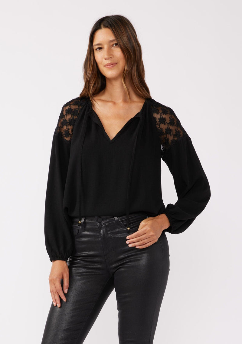 [Color: Black] A front facing image of a brunette model wearing a black bohemian fall blouse with long sleeves, a split v neckline with ties, and sheer embroidered mesh detail along the shoulders.