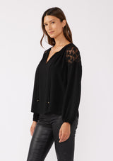 [Color: Black] A side facing image of a brunette model wearing a black bohemian fall blouse with long sleeves, a split v neckline with ties, and sheer embroidered mesh detail along the shoulders.