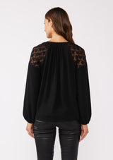 [Color: Black] A back facing image of a brunette model wearing a black bohemian fall blouse with long sleeves, a split v neckline with ties, and sheer embroidered mesh detail along the shoulders.