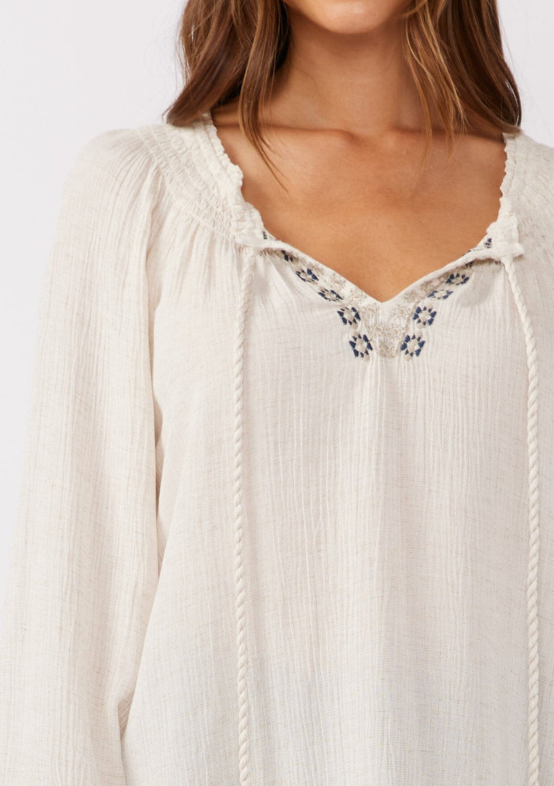 [Color: Natural] A detailed image of a brunette model wearing a bohemian blouse with embroidered details. With voluminous long sleeves, a split v neckline with tassel ties, a smocked neckline, and a relaxed fit.