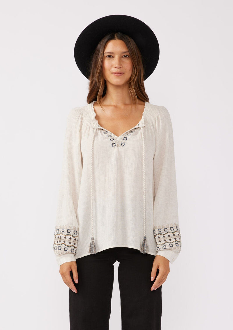 [Color: Natural] A front facing image of a brunette model wearing a bohemian blouse with embroidered details. With voluminous long sleeves, a split v neckline with tassel ties, a smocked neckline, and a relaxed fit.