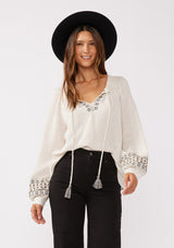 [Color: Natural] A front facing image of a brunette model wearing a bohemian blouse with embroidered details. With voluminous long sleeves, a split v neckline with tassel ties, a smocked neckline, and a relaxed fit.