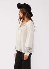 [Color: Natural] A side facing image of a brunette model wearing a bohemian blouse with embroidered details. With voluminous long sleeves, a split v neckline with tassel ties, a smocked neckline, and a relaxed fit.