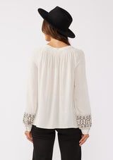 [Color: Natural] A back facing image of a brunette model wearing a bohemian blouse with embroidered details. With voluminous long sleeves, a split v neckline with tassel ties, a smocked neckline, and a relaxed fit.