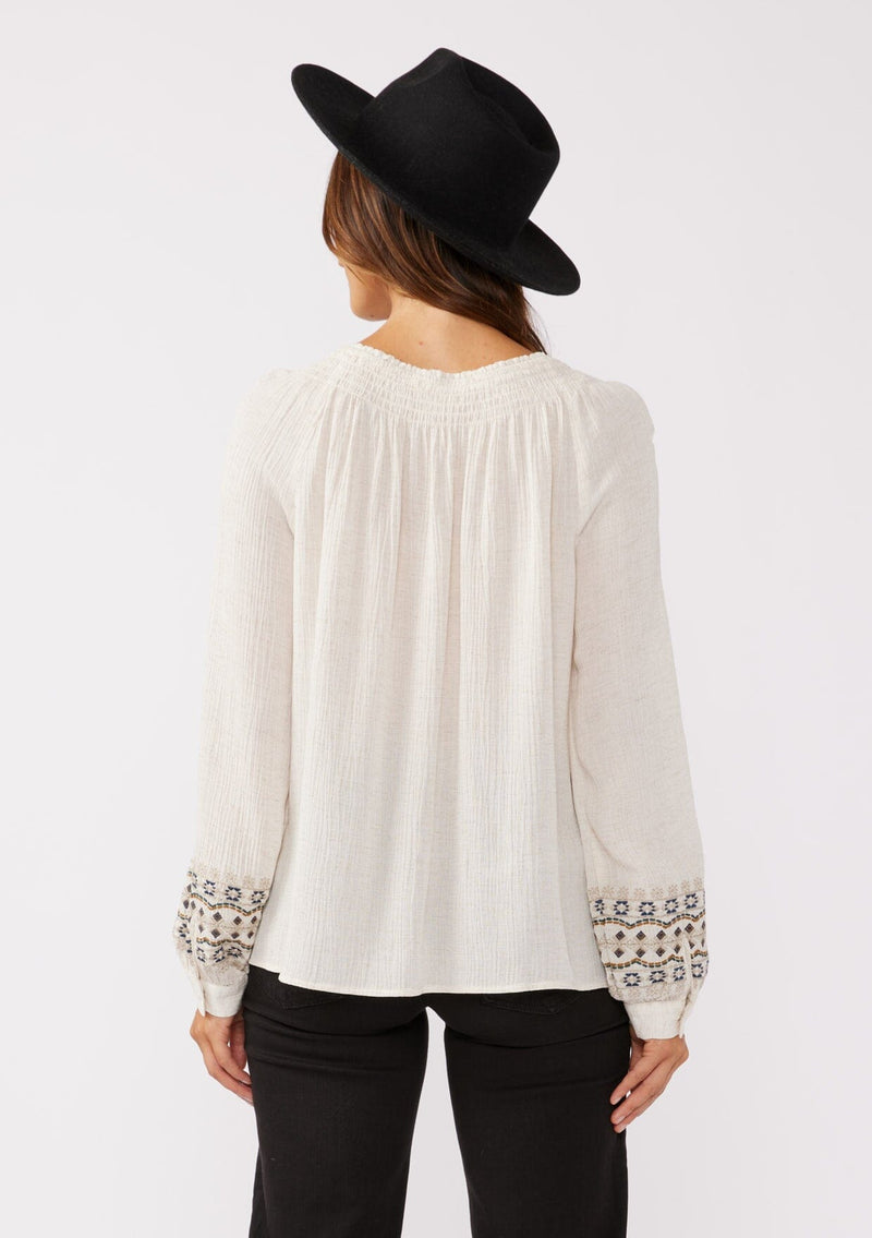 [Color: Natural] A back facing image of a brunette model wearing a bohemian blouse with embroidered details. With voluminous long sleeves, a split v neckline with tassel ties, a smocked neckline, and a relaxed fit.