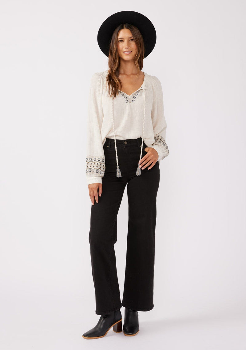 [Color: Natural] A front facing image of a brunette model wearing a bohemian blouse with embroidered details. With voluminous long sleeves, a split v neckline with tassel ties, a smocked neckline, and a relaxed fit.