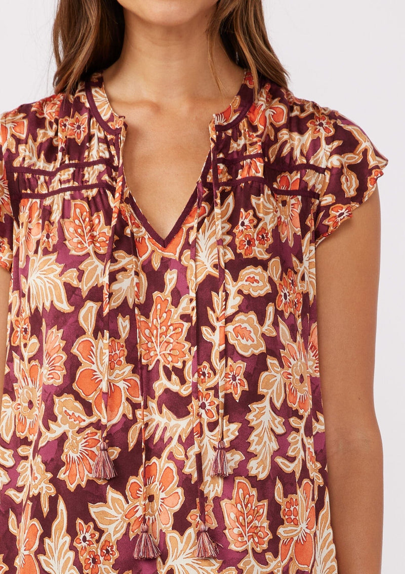 [Color: Wine/Dusty Rose] A detailed image of a brunette model wearing a red and orange bohemian floral print blouse. This autumn-inspired blouse features a double tassel tie front, short flutter sleeves, and a relaxed fit. 