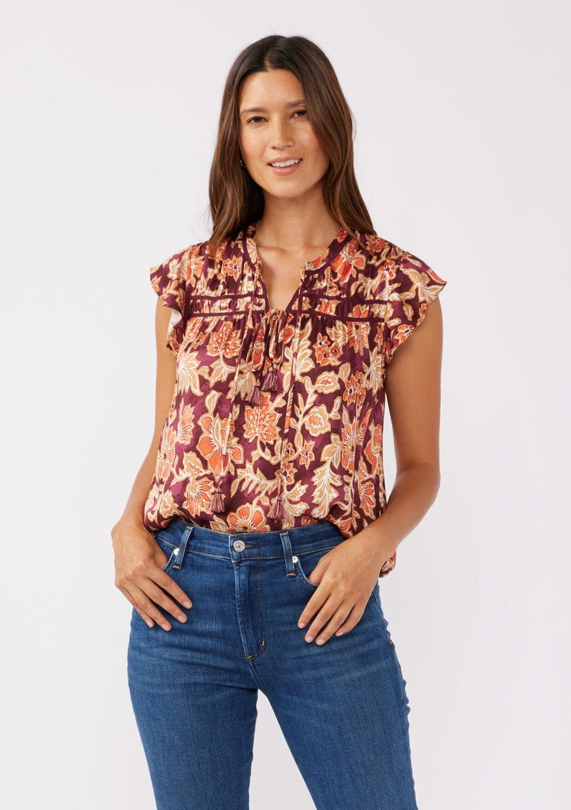 [Color: Wine/Dusty Rose] A front facing image of a brunette model wearing a red and orange bohemian floral print blouse. This autumn-inspired blouse features a double tassel tie front, short flutter sleeves, and a relaxed fit. 