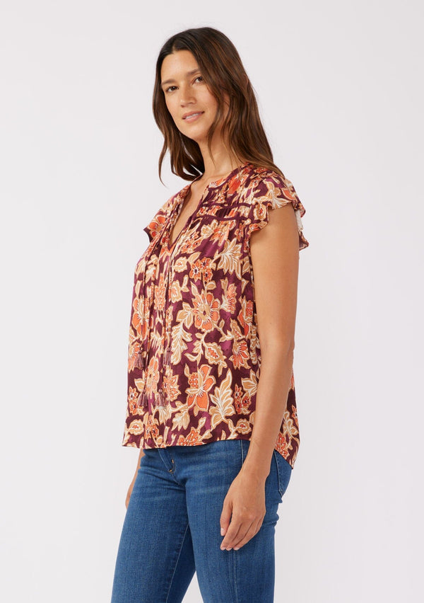 [Color: Wine/Dusty Rose] A side facing image of a brunette model wearing a red and orange bohemian floral print blouse. This autumn-inspired blouse features a double tassel tie front, short flutter sleeves, and a relaxed fit. 