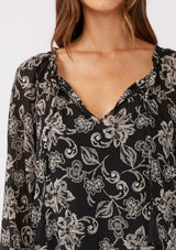 [Color: Black/Natural] A detailed image of a brunette model wearing a sheer chiffon bohemian blouse in a black and natural floral print. With long sleeves, a flowy silhouette, and a split v neckline with tassel neck ties.