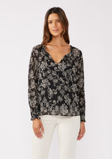 [Color: Black/Natural] A front facing image of a brunette model wearing a sheer chiffon bohemian blouse in a black and natural floral print. With long sleeves, a flowy silhouette, and a split v neckline with tassel neck ties.