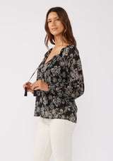 [Color: Black/Natural] A side facing image of a brunette model wearing a sheer chiffon bohemian blouse in a black and natural floral print. With long sleeves, a flowy silhouette, and a split v neckline with tassel neck ties.