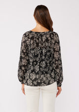 [Color: Black/Natural] A back facing image of a brunette model wearing a sheer chiffon bohemian blouse in a black and natural floral print. With long sleeves, a flowy silhouette, and a split v neckline with tassel neck ties.