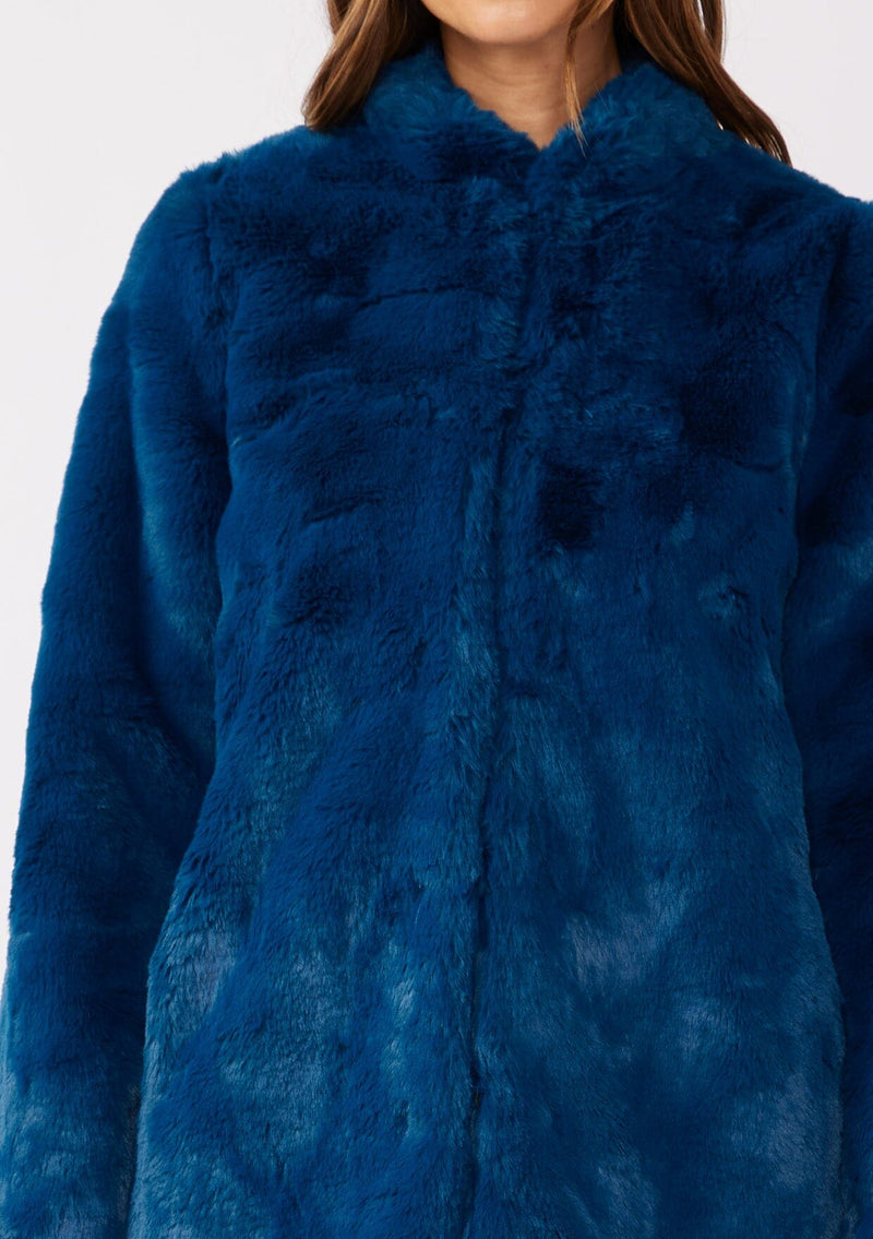 [Color: Electric Teal] A brunette model wearing an ultra luxe faux fur statement coat in bright bold blue color. Featuring essential side pockets, a silky lining, and a soft hand feel. Perfect for fall and winter when the temperatures drop. Styled with a simple black mini dress and heels. 