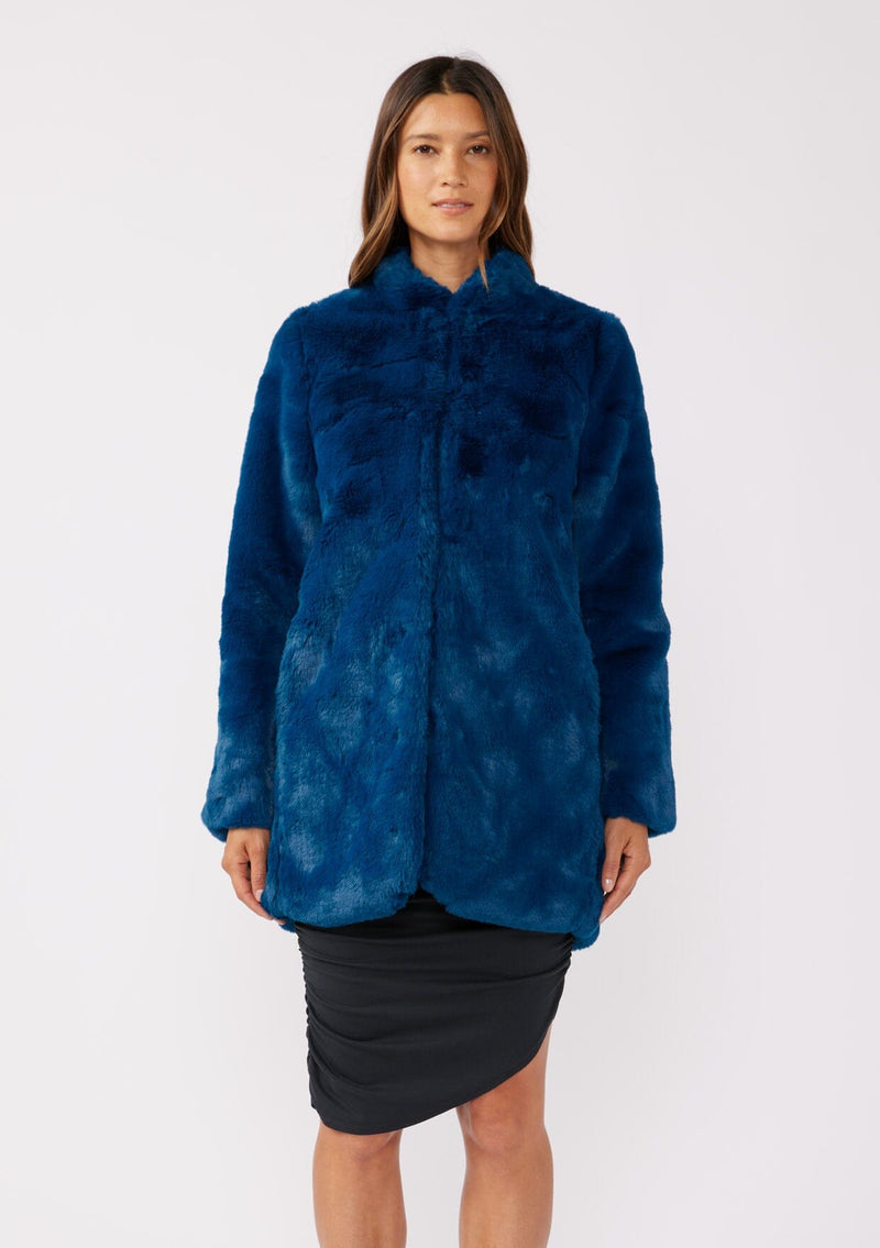 [Color: Electric Teal] A brunette model wearing an ultra luxe faux fur statement coat in bright bold blue color. Featuring essential side pockets, a silky lining, and a soft hand feel. Perfect for fall and winter when the temperatures drop. Styled with a simple black mini dress and heels. 