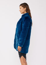 [Color: Electric Teal] A brunette model wearing an ultra luxe faux fur statement coat in bright bold blue color. Featuring essential side pockets, a silky lining, and a soft hand feel. Perfect for fall and winter when the temperatures drop. Styled with a simple black mini dress and heels. 