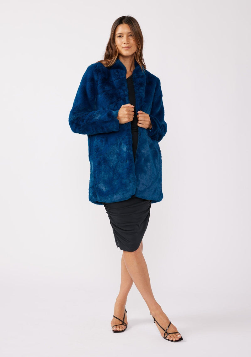 [Color: Electric Teal] A brunette model wearing an ultra luxe faux fur statement coat in bright bold blue color. Featuring essential side pockets, a silky lining, and a soft hand feel. Perfect for fall and winter when the temperatures drop. Styled with a simple black mini dress and heels. 