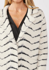 [Color: Cream/Black] A detail image of a blonde model wearing a soft and fuzzy sweater coat in a white and black chevron design. With a snap button front, side pockets, and a classic notched lapel.