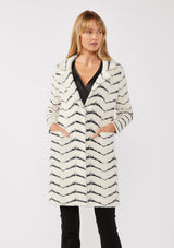 [Color: Cream/Black] A front facing image of a blonde model wearing a soft and fuzzy sweater coat in a white and black chevron design. With a snap button front, side pockets, and a classic notched lapel.