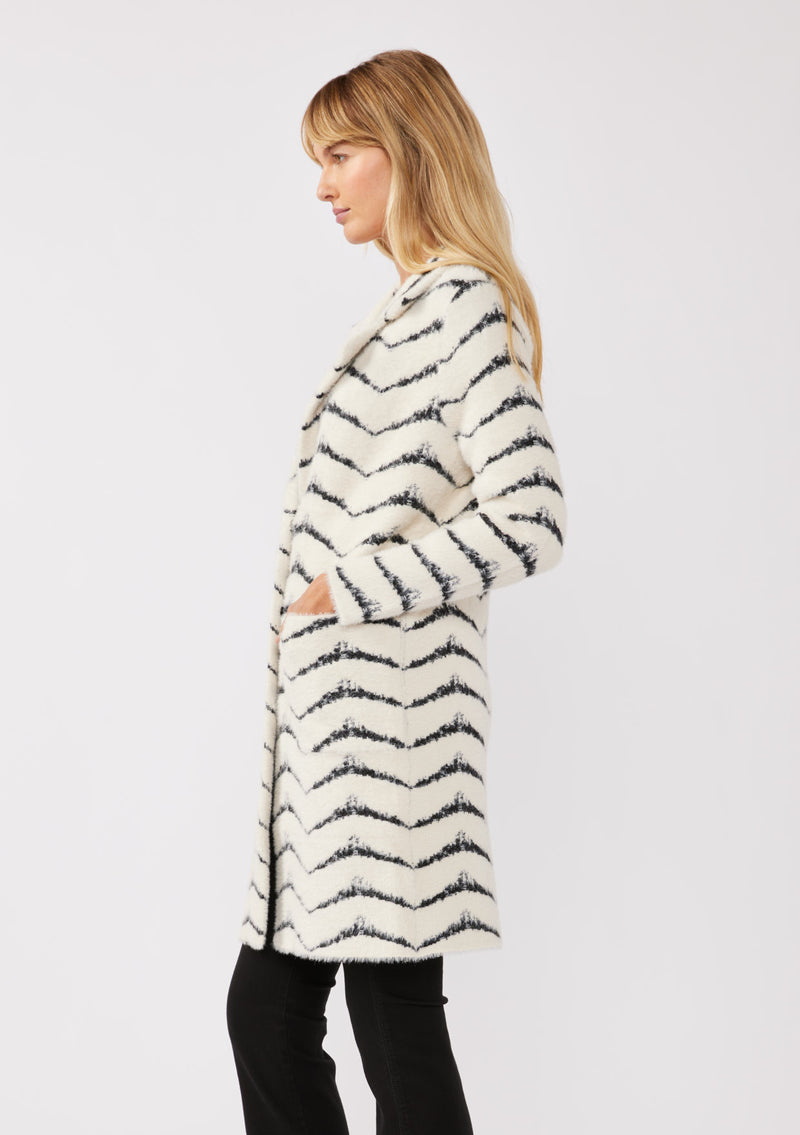 [Color: Cream/Black] A side facing image of a blonde model wearing a soft and fuzzy sweater coat in a white and black chevron design. With a snap button front, side pockets, and a classic notched lapel.