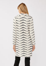 [Color: Cream/Black] A back facing image of a blonde model wearing a soft and fuzzy sweater coat in a white and black chevron design. With a snap button front, side pockets, and a classic notched lapel.