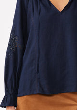 [Color: Navy] A detail image of a blonde model wearing a bohemian navy blue blouse. With a split v neckline, long sleeves, ruffled elastic wrist cuffs, and embroidered lace details. A fall blouse styled with trendy suede skirt. 