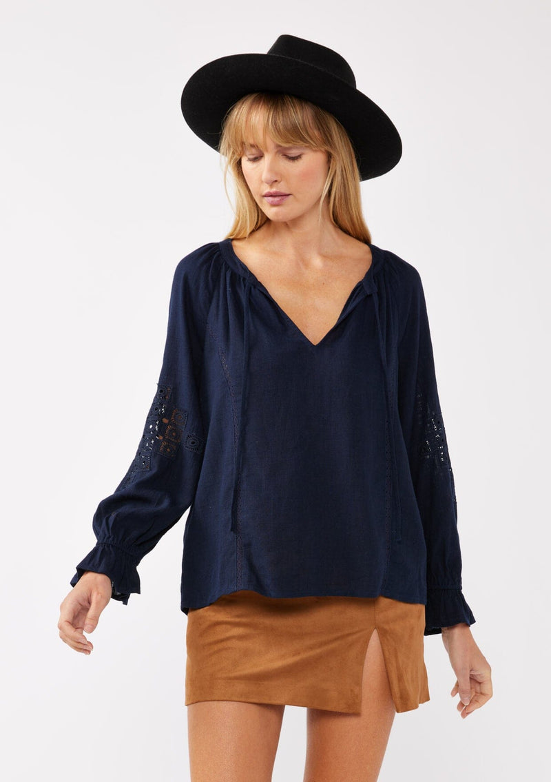 [Color: Navy] A front facing image of a blonde model wearing a bohemian navy blue blouse. With a split v neckline, long sleeves, ruffled elastic wrist cuffs, and embroidered lace details. A fall blouse styled with trendy suede skirt. 