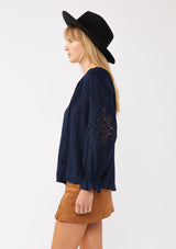 [Color: Navy] A side facing image of a blonde model wearing a bohemian navy blue blouse. With a split v neckline, long sleeves, ruffled elastic wrist cuffs, and embroidered lace details. A fall blouse styled with trendy suede skirt. 