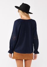 [Color: Navy] A back facing image of a blonde model wearing a bohemian navy blue blouse. With a split v neckline, long sleeves, ruffled elastic wrist cuffs, and embroidered lace details. A fall blouse styled with trendy suede skirt. 