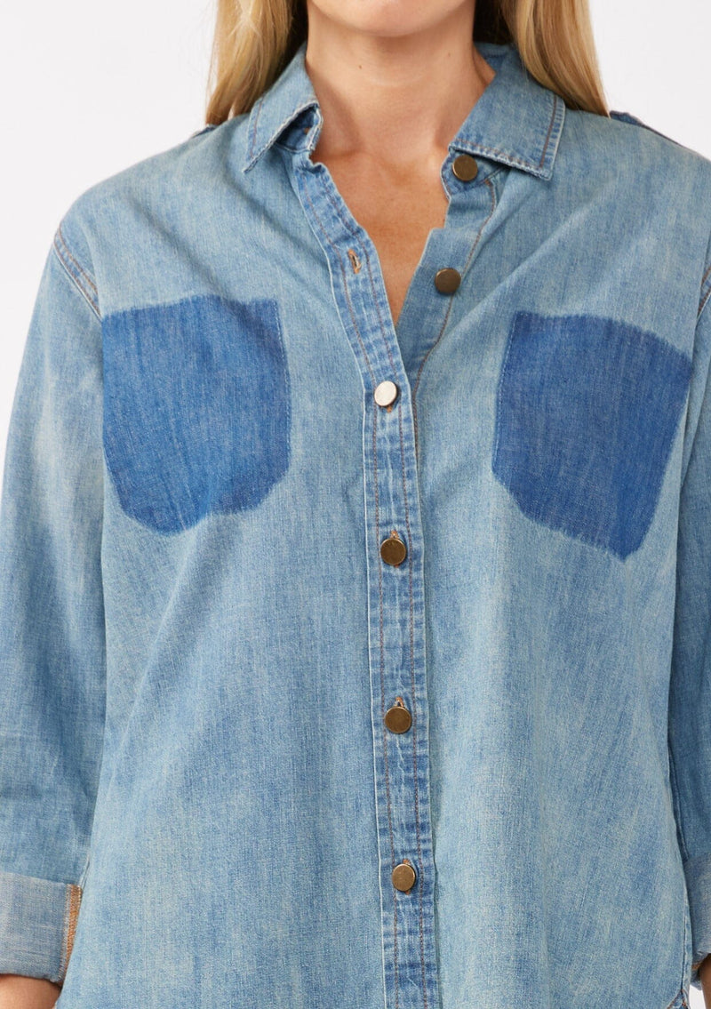 [Color: Indigo] A detail image of a blonde model wearing a two-tone cotton button-down shirt in denim indigo. The blouse features a collared neckline, faux pockets, and roll-tab sleeves, offering a western-inspired style perfect for fall looks.