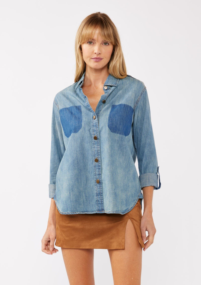 [Color: Indigo] A front facing image of a blonde model wearing a two-tone cotton button-down shirt in denim indigo. The blouse features a collared neckline, faux pockets, and roll-tab sleeves, offering a western-inspired style perfect for fall looks.