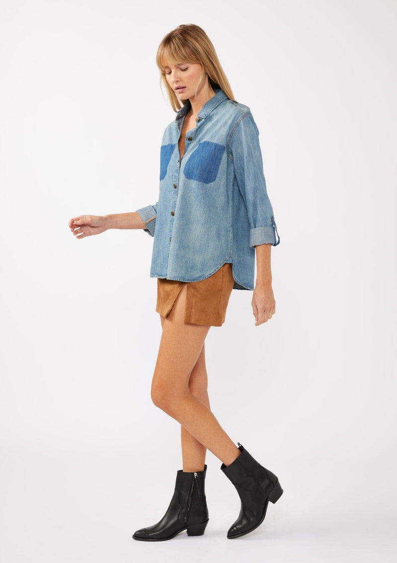 [Color: Indigo] An image of a blonde model wearing a two-tone cotton button-down shirt in denim indigo. The blouse features a collared neckline, faux pockets, and roll-tab sleeves, offering a western-inspired style perfect for fall looks.