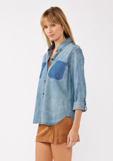 [Color: Indigo] A side facing image of a blonde model wearing a two-tone cotton button-down shirt in denim indigo. The blouse features a collared neckline, faux pockets, and roll-tab sleeves, offering a western-inspired style perfect for fall looks.