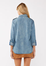 [Color: Indigo] A back facing image of a blonde model wearing a two-tone cotton button-down shirt in denim indigo. The blouse features a collared neckline, faux pockets, and roll-tab sleeves, offering a western-inspired style perfect for fall looks.