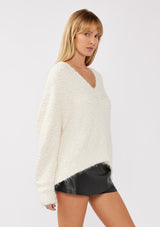 [Color: Antique White] A side-facing image of a blonde model wearing a green cozy, fuzzy popcorn-textured pullover sweater with long sleeves, a classic V-neckline, and a high-low hemline. Ideal for lounging or casual fall outings.