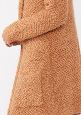 [Color: Camel] A detail image of a blonde model wearing a fuzzy brown fall cardigan with a popcorn texture. The cardigan features an open front, large cozy patch pockets, and an attached hoodie, draped for warmth and ideal for layering. 