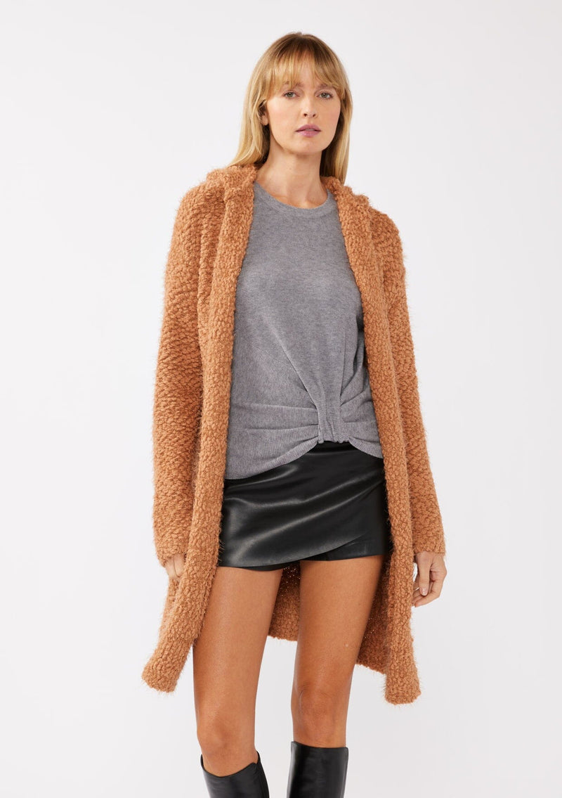 [Color: Camel] A front facing image of a blonde model wearing a fuzzy brown fall cardigan with a popcorn texture. The cardigan features an open front, large cozy patch pockets, and an attached hoodie, draped for warmth and ideal for layering.