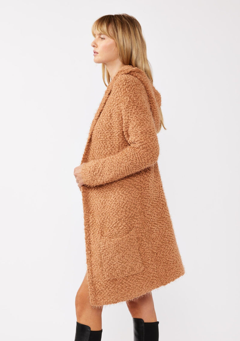 [Color: Camel] A side facing image of a blonde model wearing a fuzzy brown fall cardigan with a popcorn texture. The cardigan features an open front, large cozy patch pockets, and an attached hoodie, draped for warmth and ideal for layering.