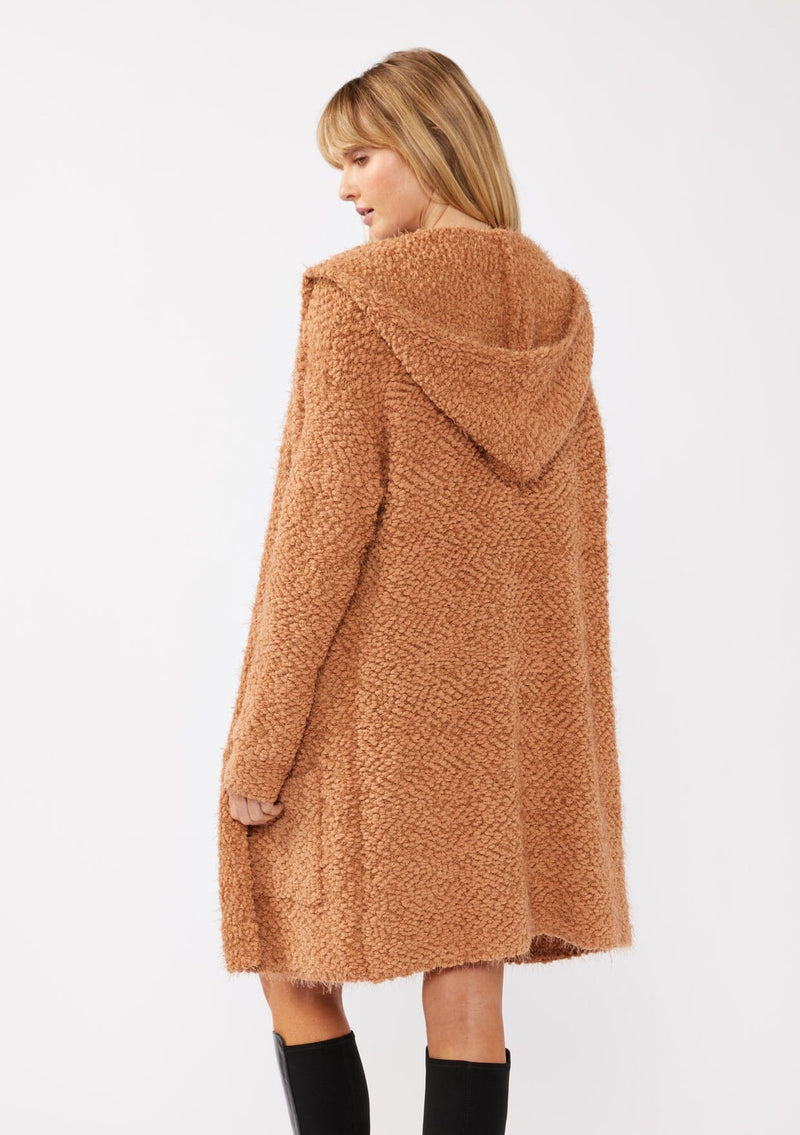 [Color: Camel] A back facing image of a blonde model wearing a fuzzy brown fall cardigan with a popcorn texture. The cardigan features an open front, large cozy patch pockets, and an attached hoodie, draped for warmth and ideal for layering.