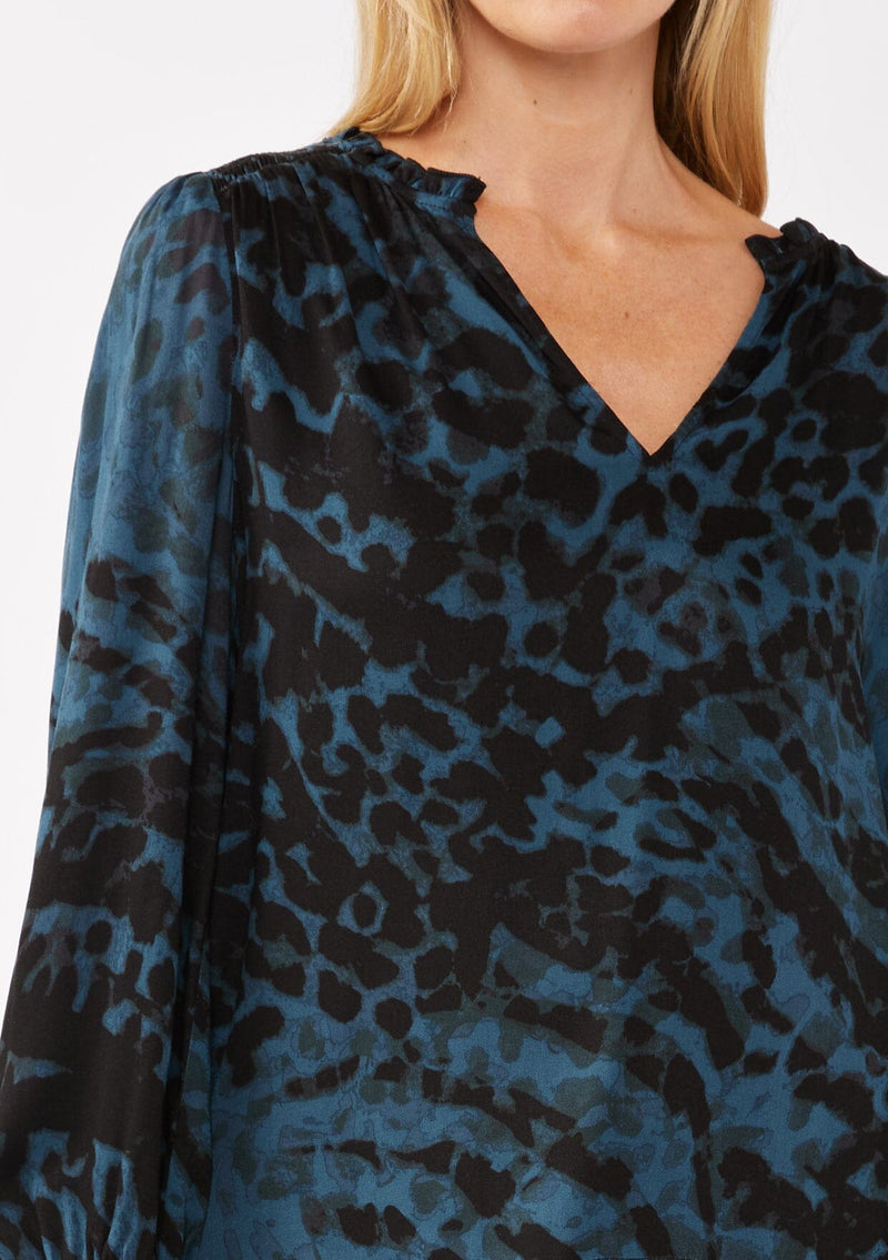 [Color: Midnight/Black] A detail image of a blonde model wearing a leopard print blouse in black and blue. With a split v-neckline, smocked cuffs, and a smocked back detail. Ultra relaxed and flowy, perfect work and special occasions. 