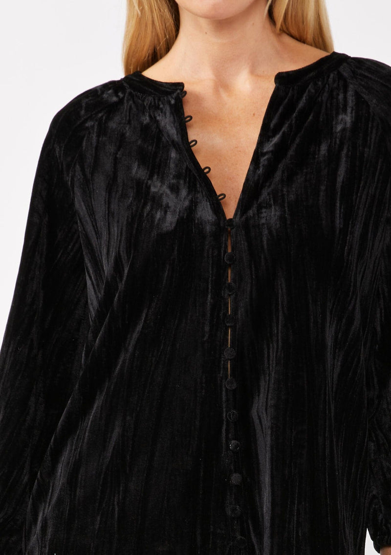 [Color: Black ] A detail image of a blonde model wearing a black velvet blouse with a loop button front, long sleeves, and elastic cuffs. The boho-inspired holiday blouse is paired with black faux leather bottoms for a chic nighttime look.