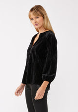 [Color: Black ] A side facing image of a blonde model wearing a black velvet blouse with a loop button front, long sleeves, and elastic cuffs. The boho-inspired holiday blouse is paired with black faux leather bottoms for a chic nighttime look.