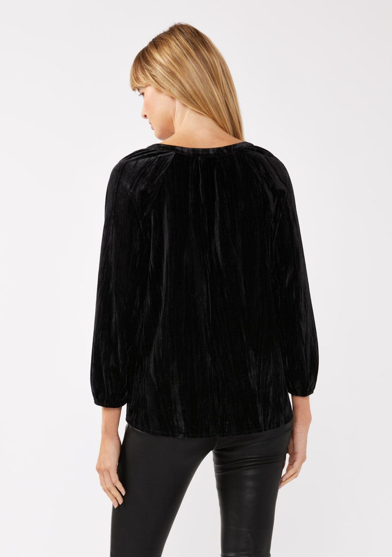 [Color: Black ] A back facing image of a blonde model wearing a black velvet blouse with a loop button front, long sleeves, and elastic cuffs. The boho-inspired holiday blouse is paired with black faux leather bottoms for a chic nighttime look.