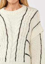 [Color: Cream/Black] A detail image of a blonde model wearing a cozy knit sweater with a mixed fabric patchwork and cable knit detailing. This casual sweater features a crew neckline, long sleeves with a dropped shoulder, and a contrasting stripe accent. A cold weather staple for the fall and winter season. 