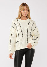 [Color: Cream/Black] A front facing image of a blonde model wearing a cozy knit sweater with a mixed fabric patchwork and cable knit detailing. This casual sweater features a crew neckline, long sleeves with a dropped shoulder, and a contrasting stripe accent. A cold weather staple for the fall and winter season. 