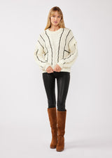 [Color: Cream/Black] A full body front facing image of a blonde model wearing a cozy knit sweater with a mixed fabric patchwork and cable knit detailing. This casual sweater features a crew neckline, long sleeves with a dropped shoulder, and a contrasting stripe accent. A cold weather staple for the fall and winter season. 