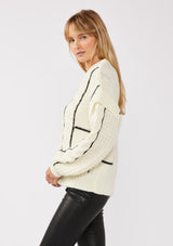 [Color: Cream/Black] A side facing image of a blonde model wearing a cozy knit sweater with a mixed fabric patchwork and cable knit detailing. This casual sweater features a crew neckline, long sleeves with a dropped shoulder, and a contrasting stripe accent. A cold weather staple for the fall and winter season. 